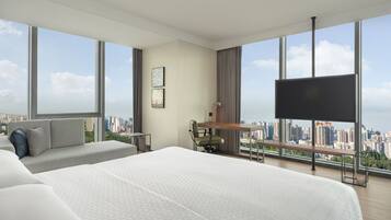 A modern hotel room with a large bed, a desk, a chair, and a TV. The room has large windows offering a city view.