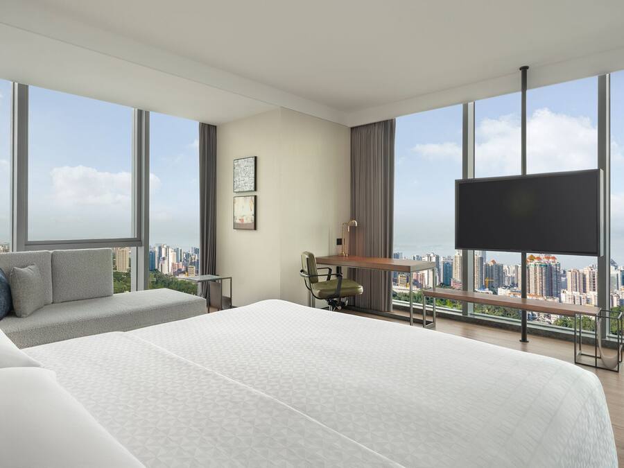 A modern hotel room with a large bed, a desk, a chair, and a TV. The room has large windows offering a city view.