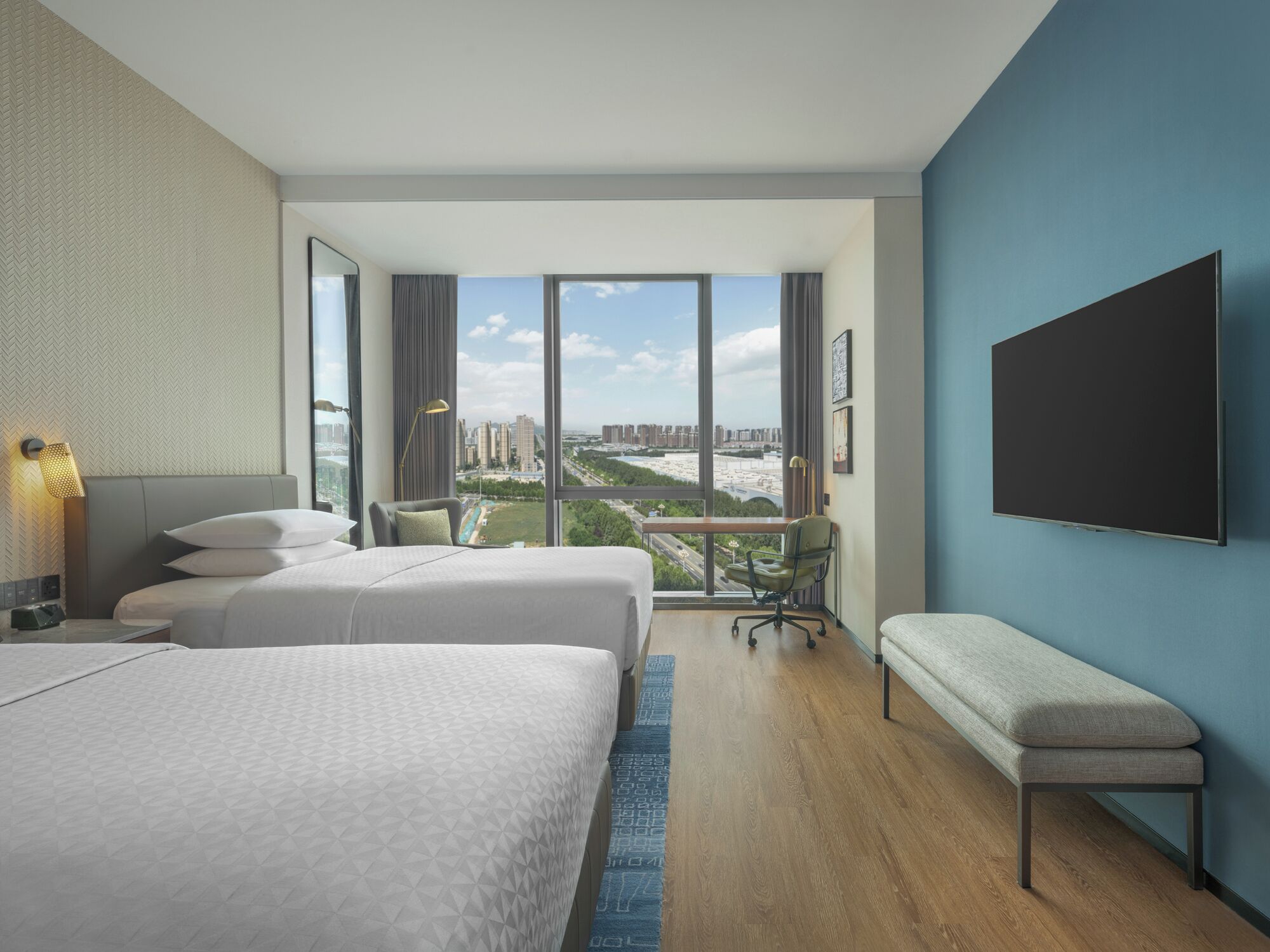 A modern hotel room with two beds, a large window offering a city view, a desk with a chair, and a mounted TV.