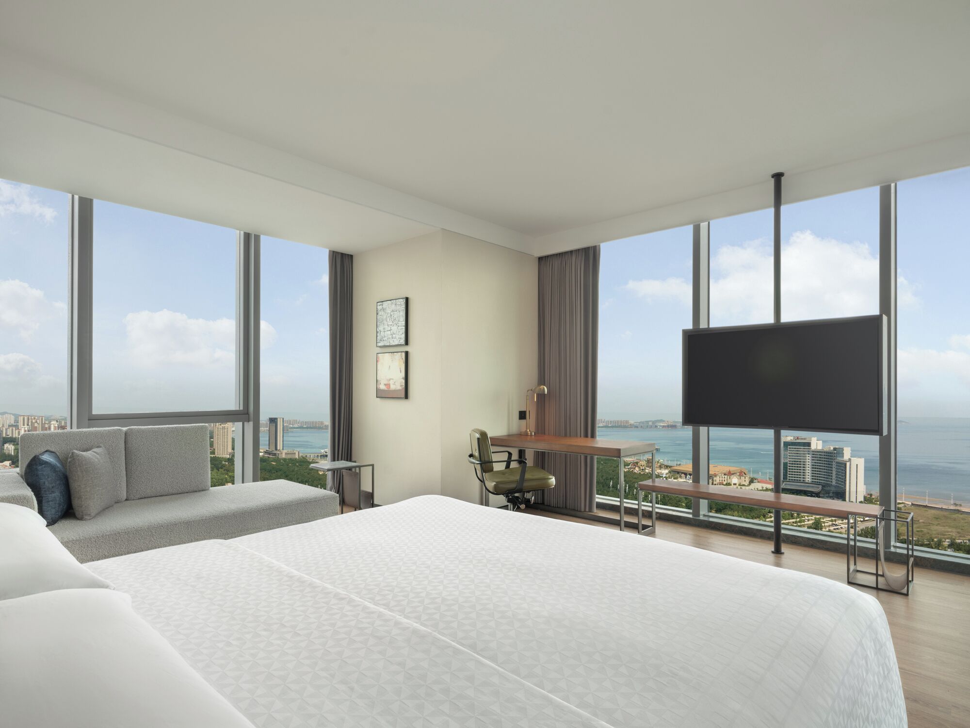 A modern hotel room with a large bed, a desk, and a TV. It has large windows with a city and ocean view.