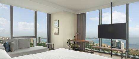 A modern hotel room with a large bed, a desk, and a TV. It has large windows with a city and ocean view.