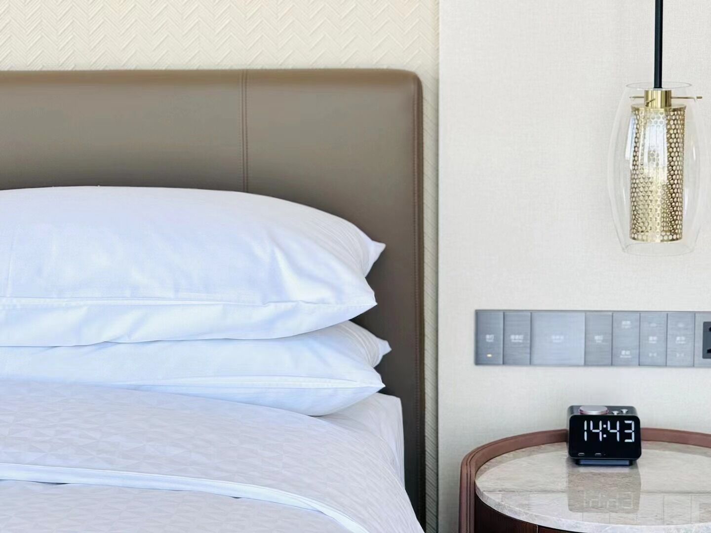 Ein ordentlich bezogenes Bett mit weißen Leinen, eine moderne Pendelleuchte, eine digitale Uhr und eine an der Wand montierte Bedieneinheit.