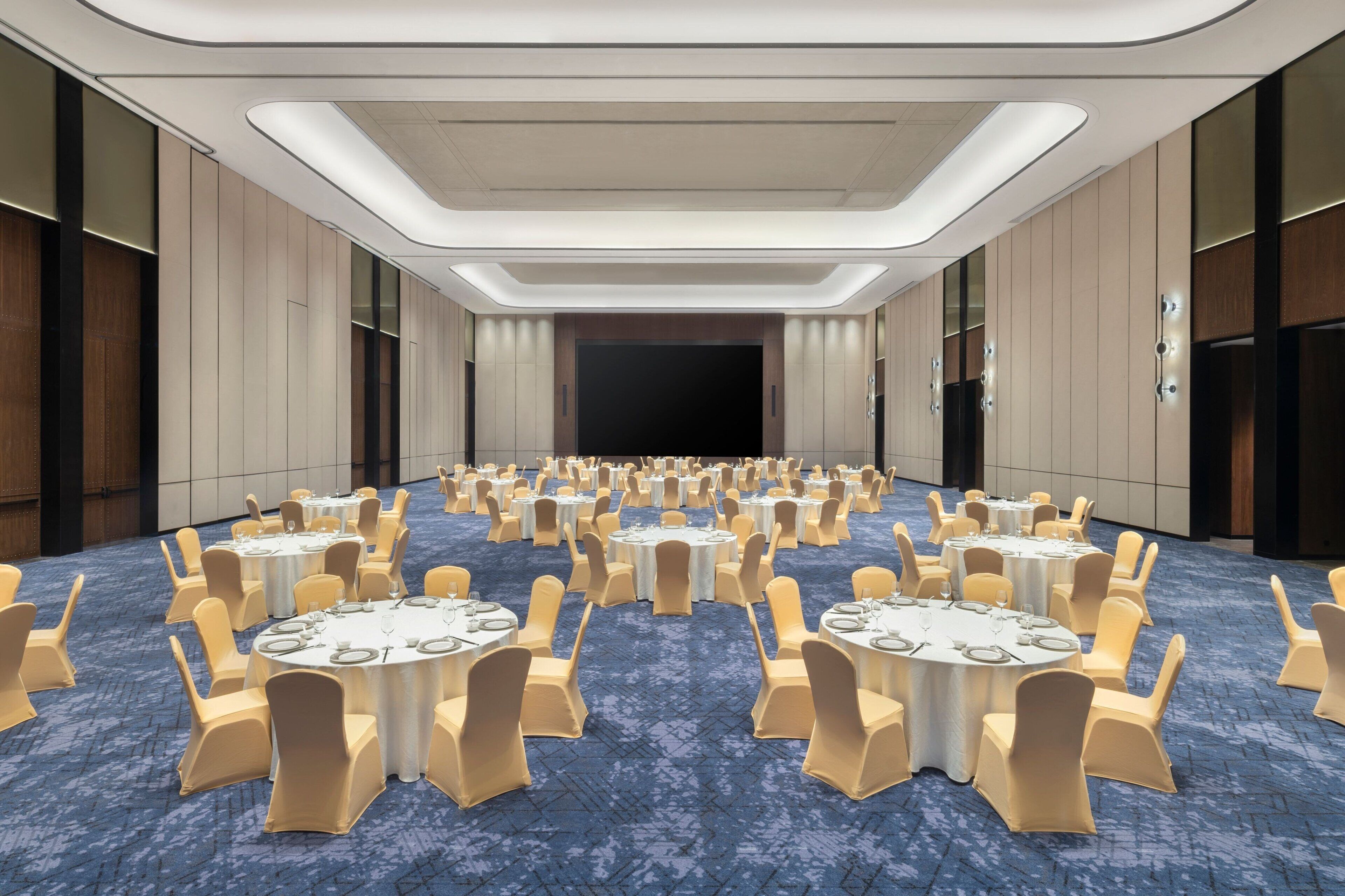 A large conference room with round tables and chairs, a stage at the front, and a carpeted floor.