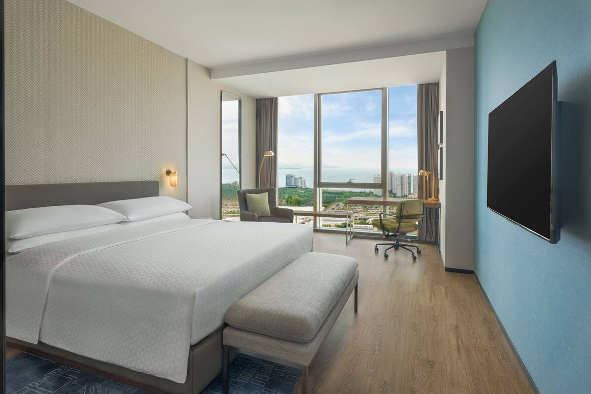 A modern hotel room with a large bed, a desk, and a view of the cityscape.