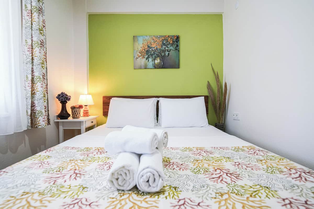 A hotel room with a bed, a nightstand with a lamp, a painting on the wall, and a window with curtains.