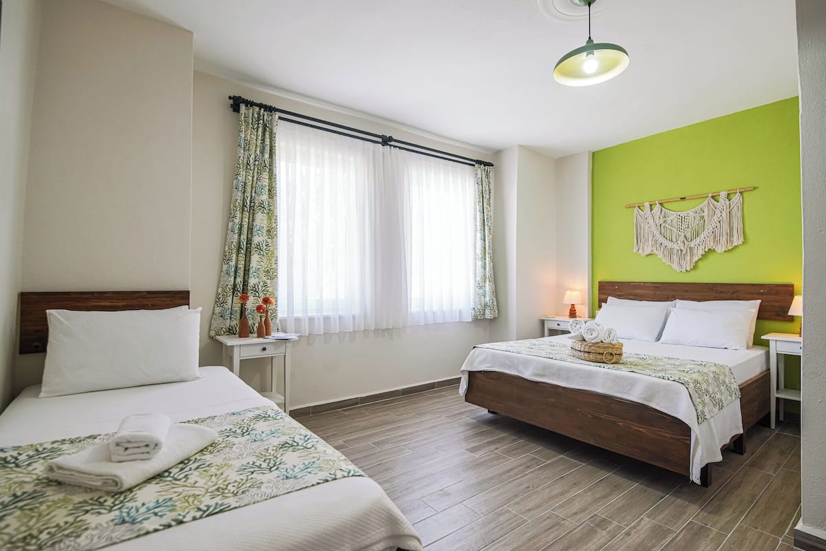 A hotel room with two beds, a green accent wall, a macrame wall hanging, and a window with sheer curtains.