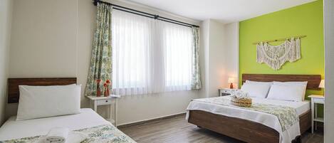 Habitación de hotel con dos camas, una pared de acento verde, un tapiz de macramé y una ventana con cortinas traslúcidas.
