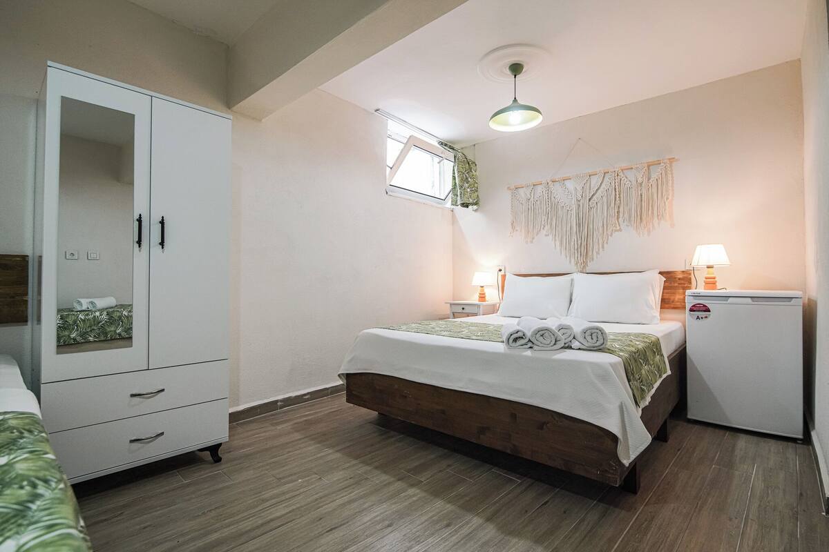 A bedroom with a bed, a wardrobe, a nightstand, a lamp, and a small refrigerator.