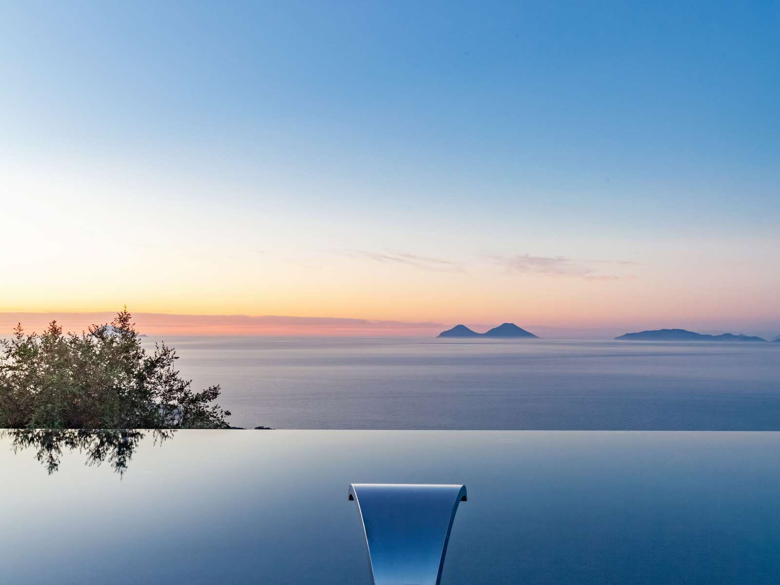An infinity pool with a clear view of the sea and distant mountains at sunset.