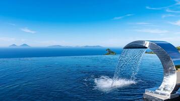 Vista panoramica su una piscina a sfioro con elemento d'acqua, circondata da un pontile in legno e affacciata su un paesaggio montuoso.