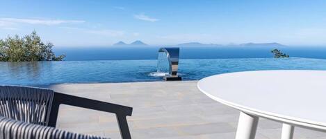 Una piscina a sfioro con vista sul mare e sulle montagne, un tavolo bianco e sedie nere.