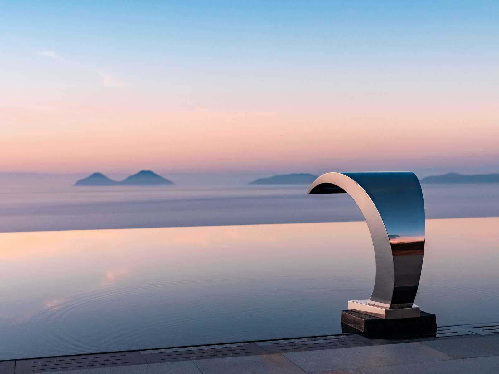 A modern, minimalist sculpture on a reflective surface with a serene landscape and distant islands in the background.
