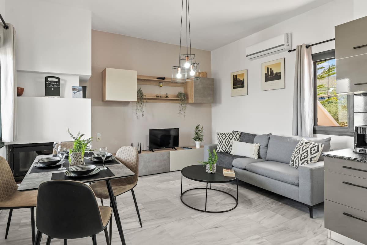 Ein modernes Wohnzimmer mit grauem Sofa, einem Esstisch mit Stühlen, einem Couchtisch und einem Fernseher. Es sind Pflanzen, eine Pendelleuchte und Kunstwerke an den Wänden zu sehen.