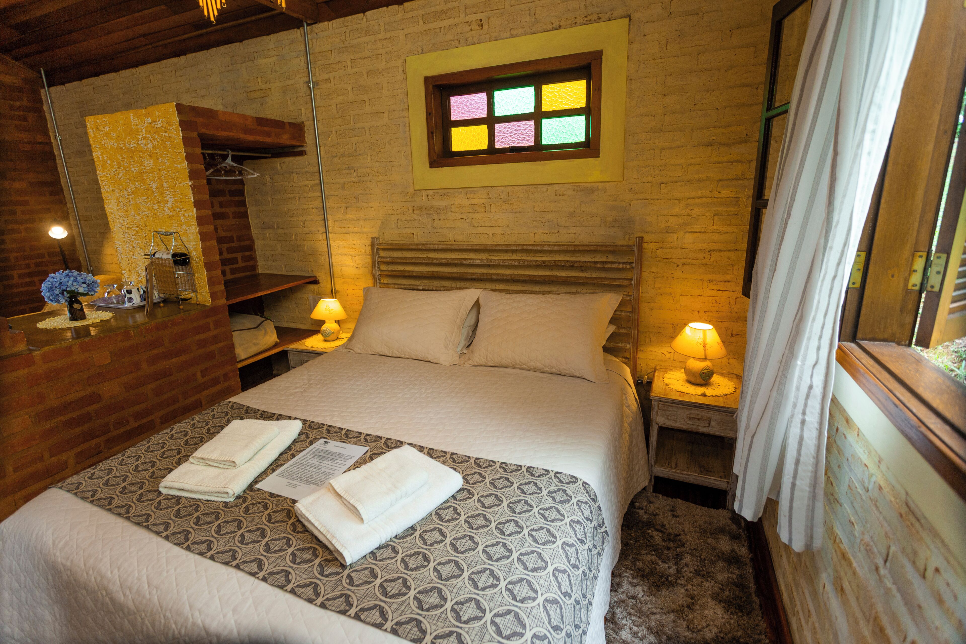 A bedroom with a patterned bedspread, a wooden headboard, a nightstand with a lamp, and a window with a stained glass design.