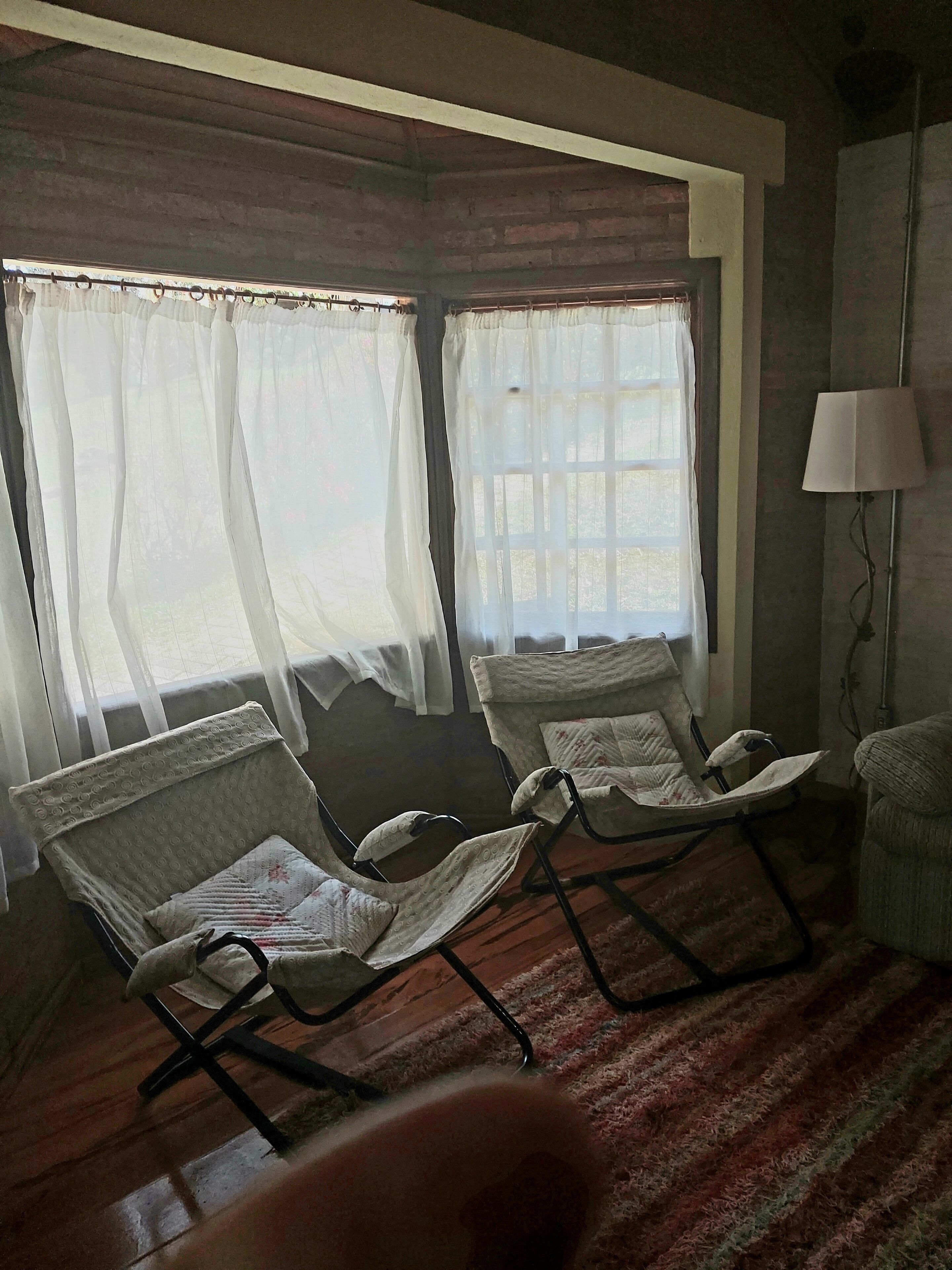 Una habitación con dos mecedoras, una lámpara y una ventana con cortinas traslúcidas.