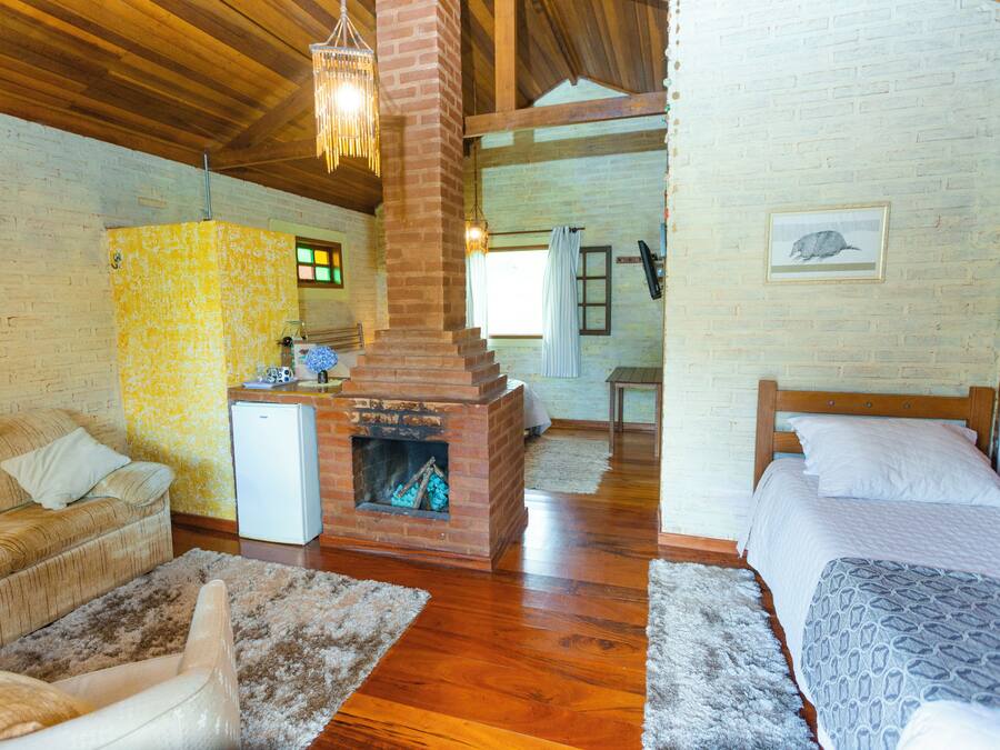 A room with a brick fireplace, wooden ceiling beams, a sofa, a bed, and a small refrigerator.