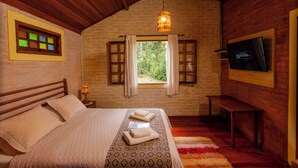 A bedroom with a wooden ceiling, a window with curtains, a bed with a patterned bedspread, a bedside lamp, and a television on the wall. - Pousada Candeias Ltda (Cunha)