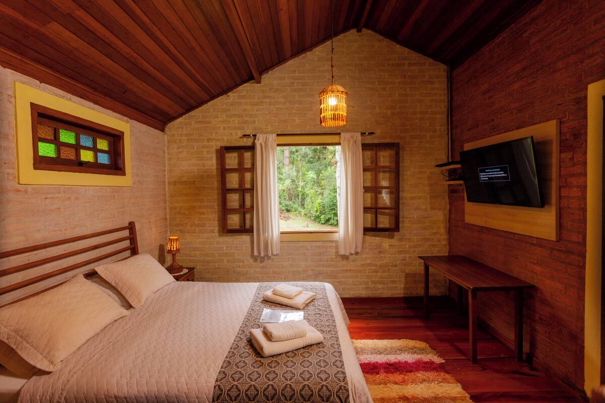 A bedroom with a wooden ceiling, a window with curtains, a bed with a patterned bedspread, a bedside lamp, and a television on the wall.