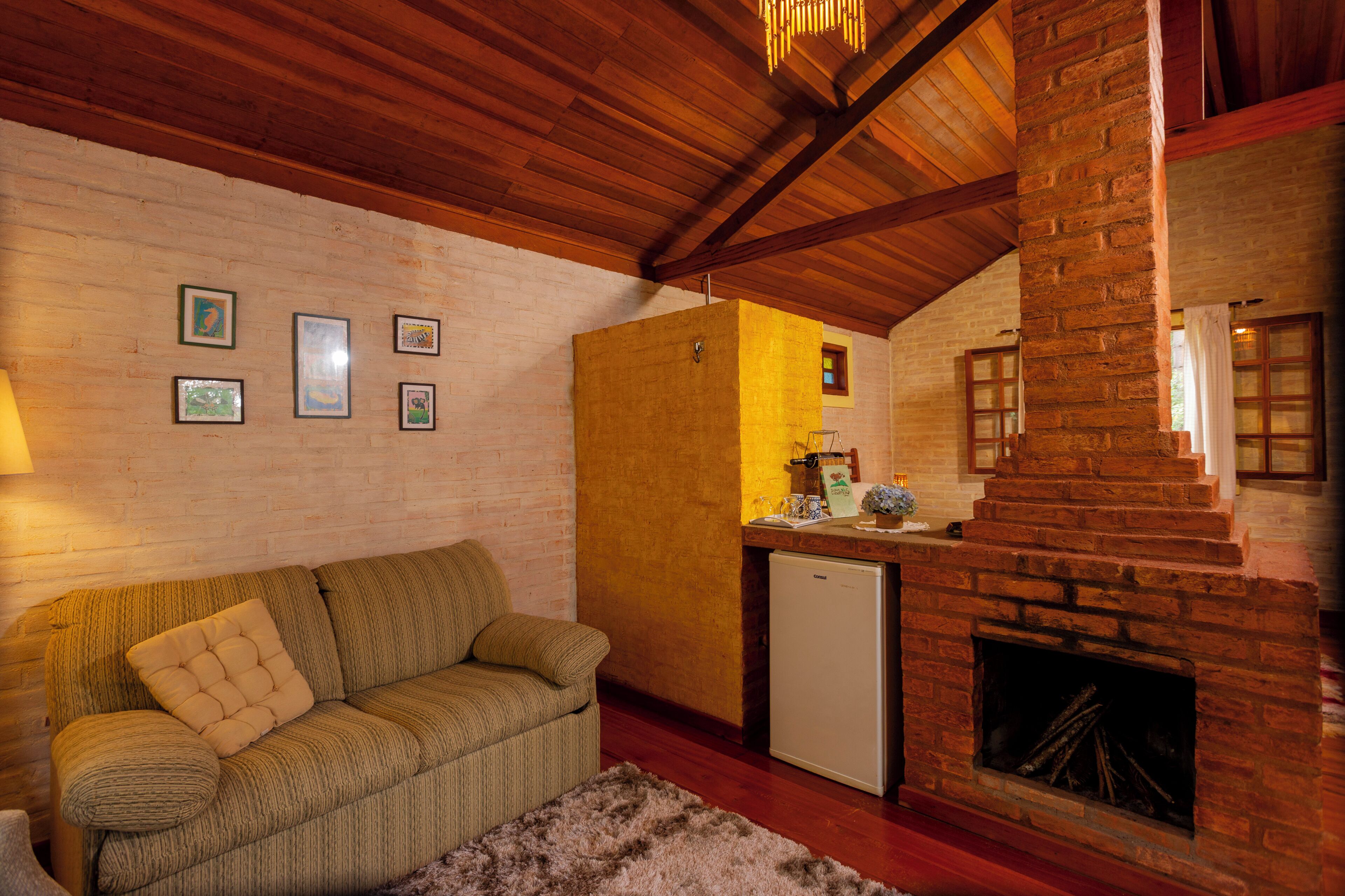 Una habitación acogedora con techo de madera, chimenea de ladrillo, un sofá, un refrigerador pequeño y cuadros enmarcados en la pared.