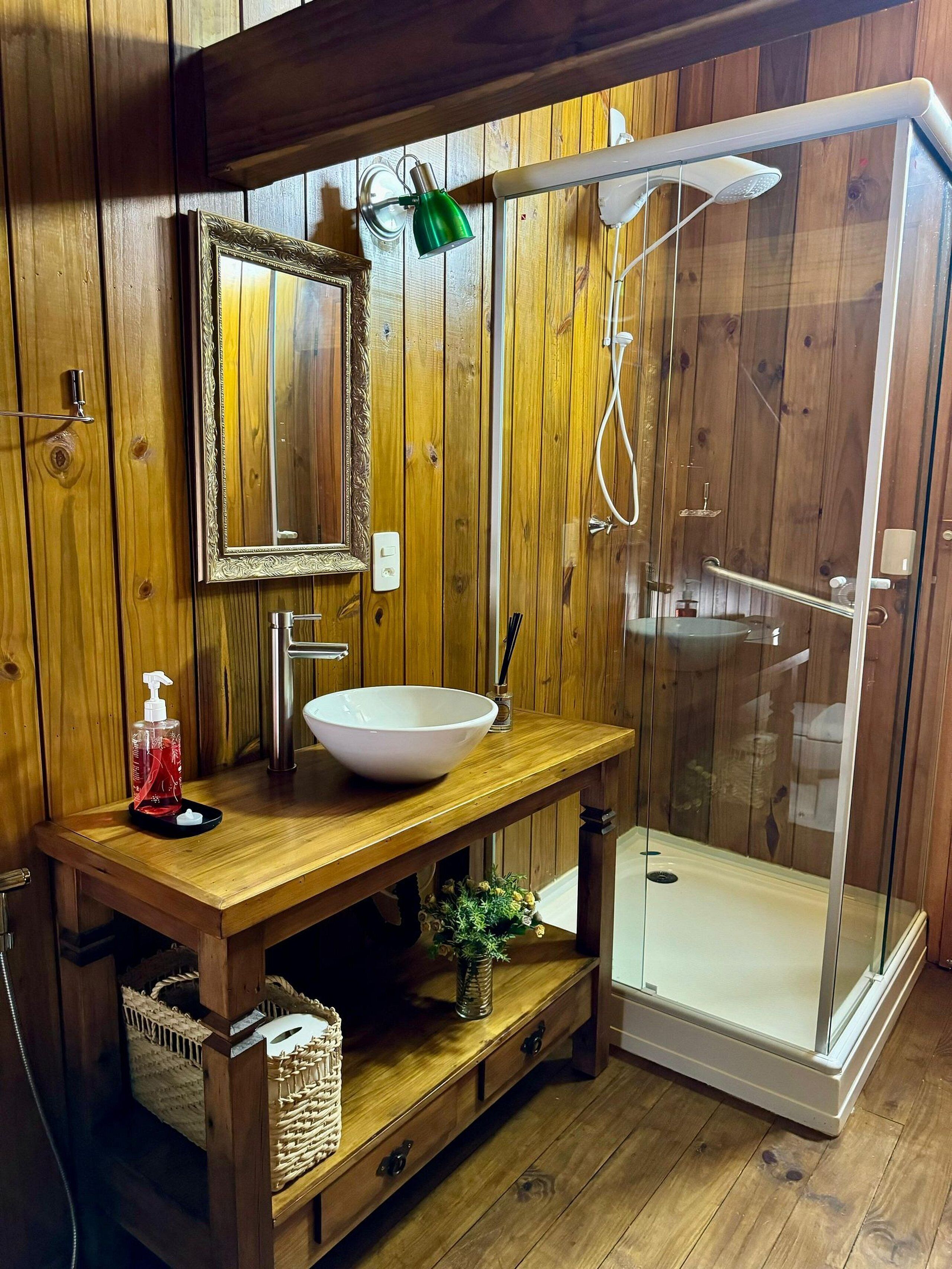 Une salle de bain en bois avec un grand miroir, un lavabo blanc, une lampe verte à main et une douche fermée par des parois de verre.