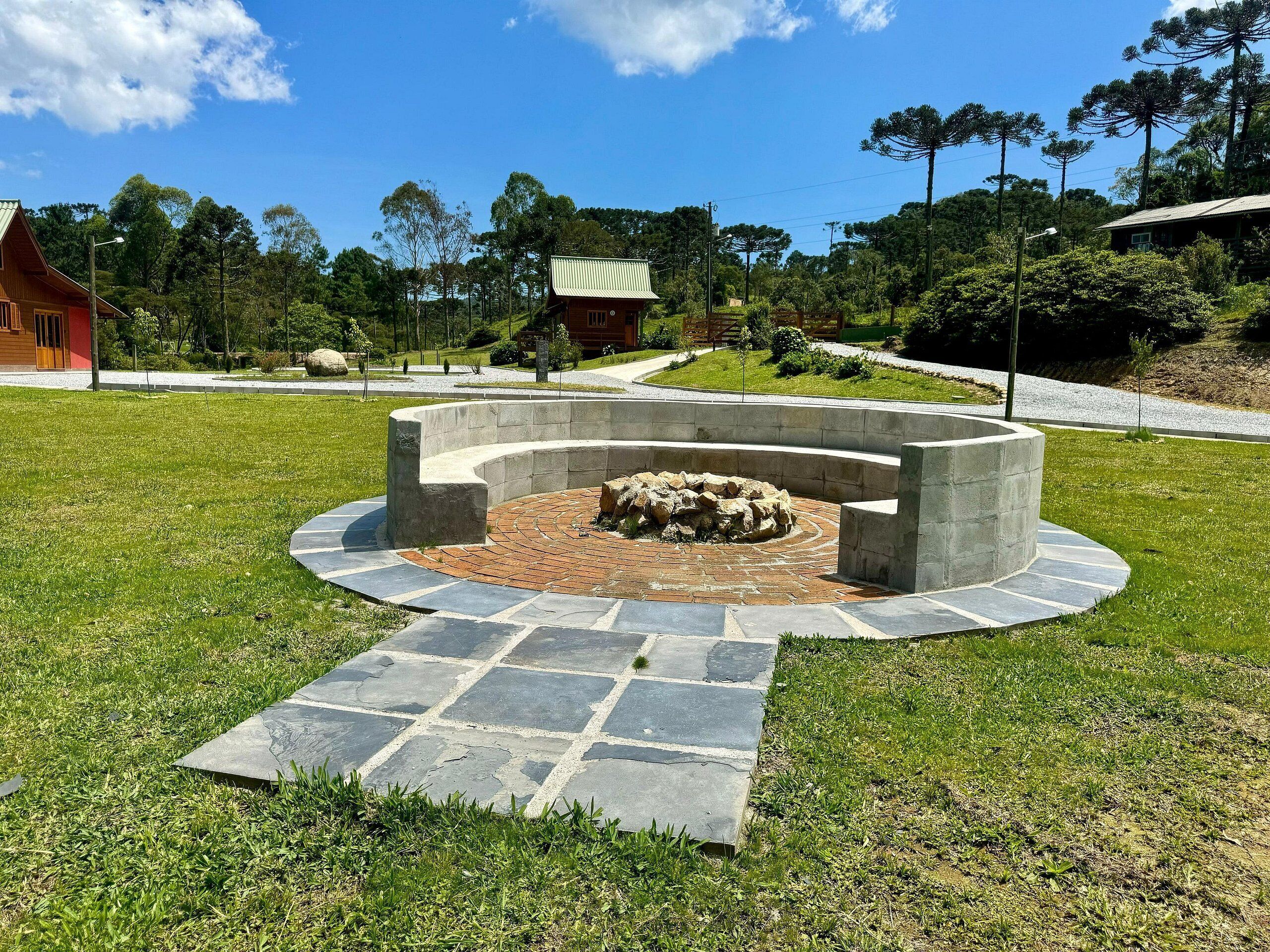 A circular stone fire pit surrounded by a paved walkway, set in a grassy area with trees and a building in the background.