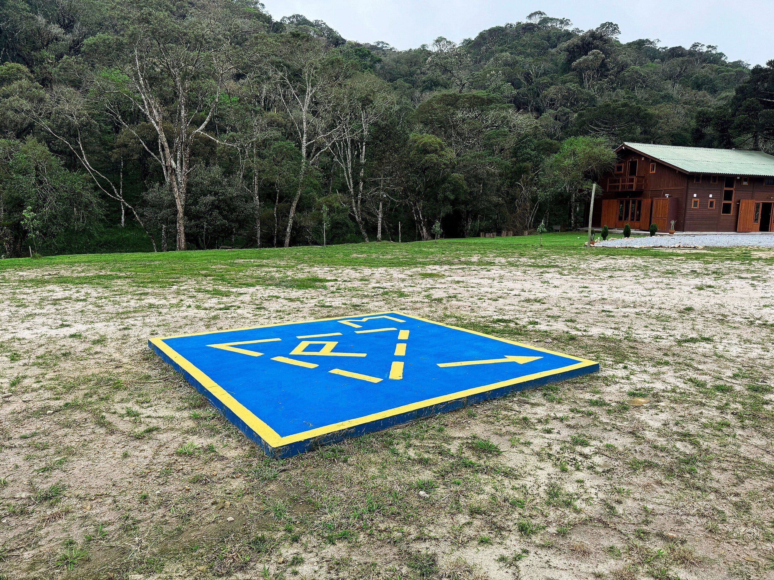 A blue and yellow directional sign on a grassy field with a forested hill in the background and a building with a green roof.
