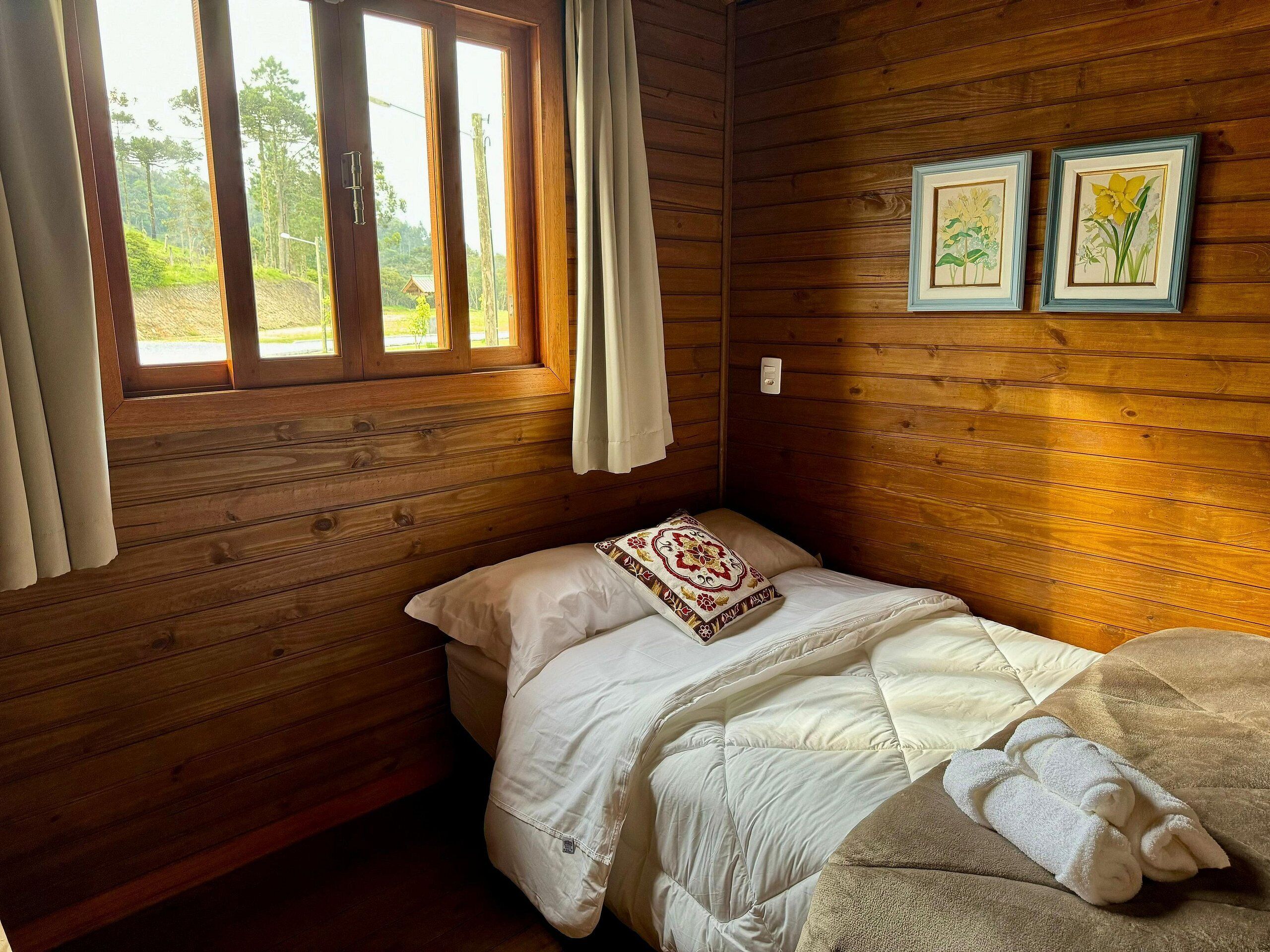Habitación de cabaña de madera con cama, ventana con cortinas e ilustraciones botánicas enmarcadas en la pared.