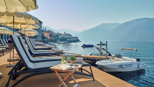 Eine Holzterrasse mit Liegestühlen und Sonnenschirmen, ein kleiner Tisch mit einem Buch und einer Pflanze sowie ein Boot, das im Hafen liegt.