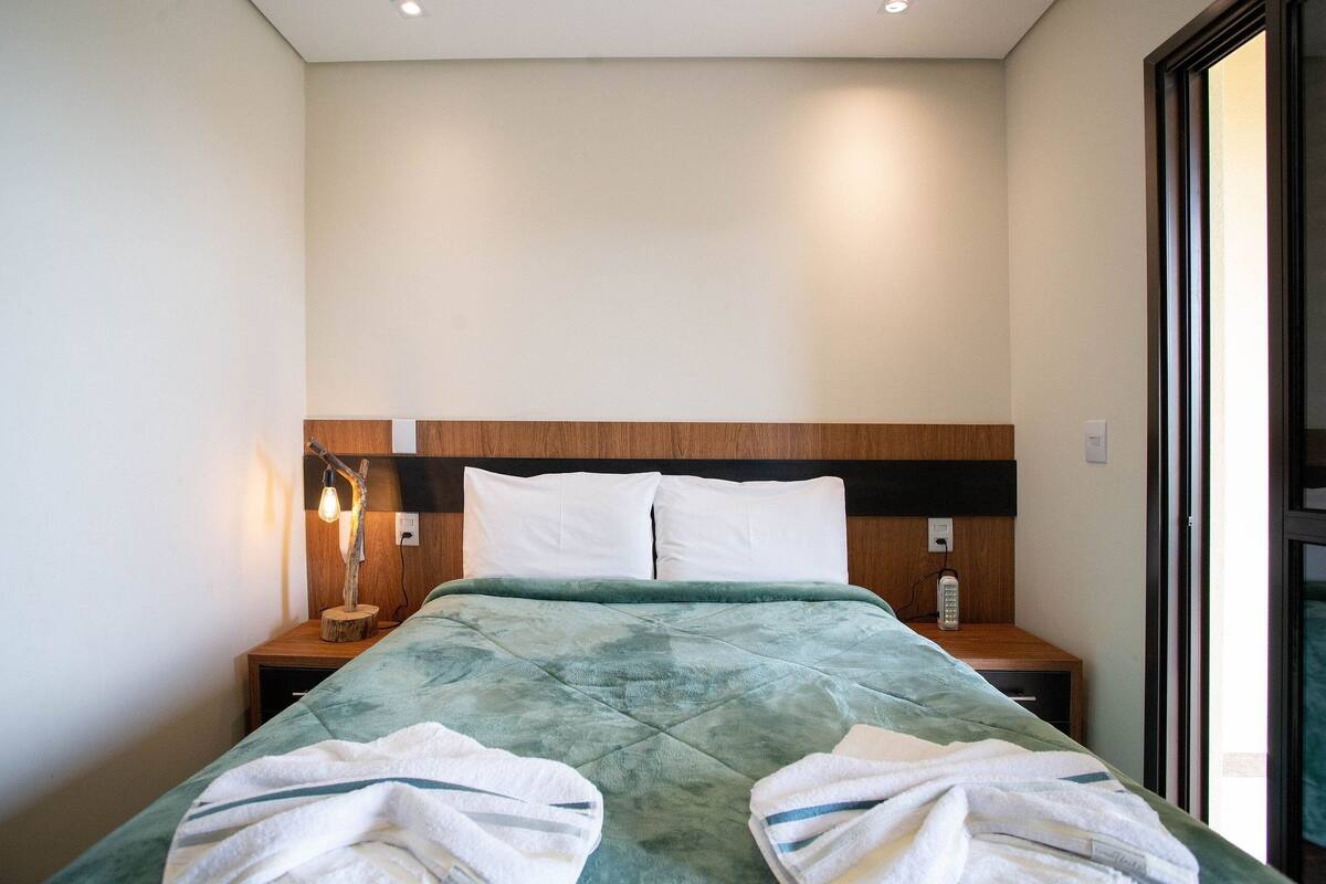 Un lit bien fait, recouvert d’une couverture verte, avec des oreillers blancs et deux tables de chevet dotées de lampes.