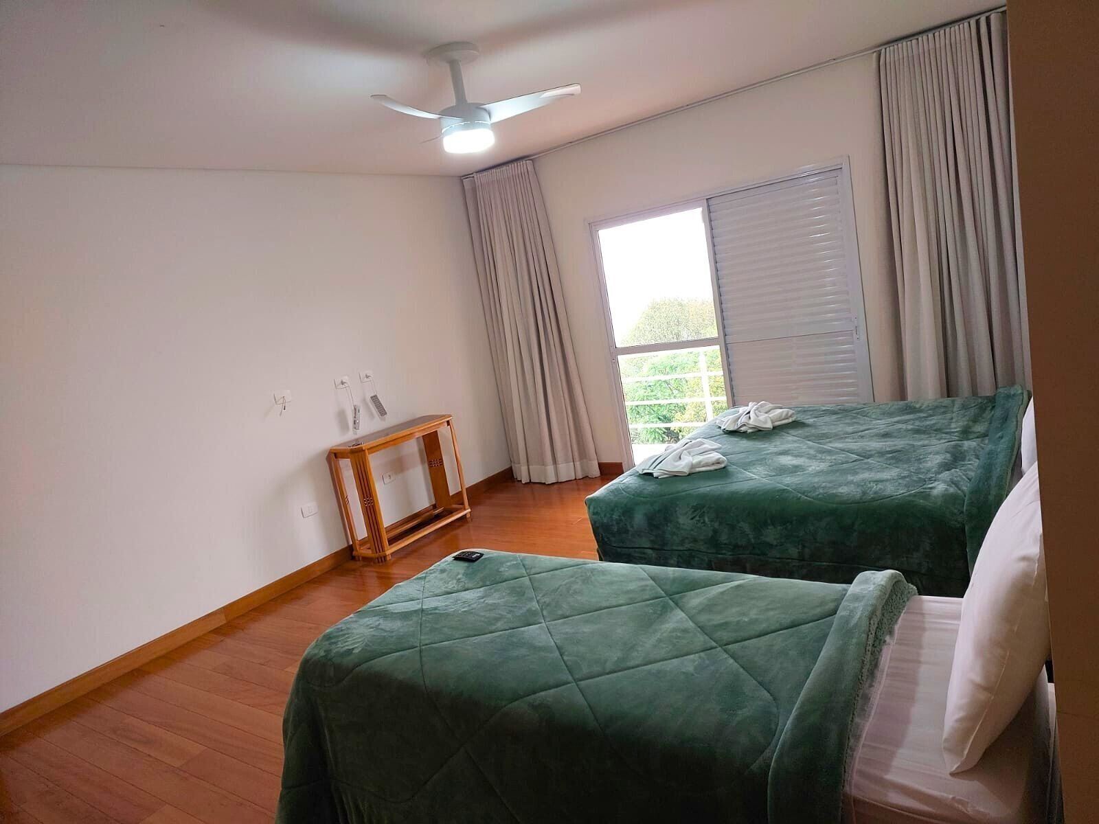 Quarto com duas camas, um ventilador de teto, uma pequena mesa de madeira e uma janela com cortinas.