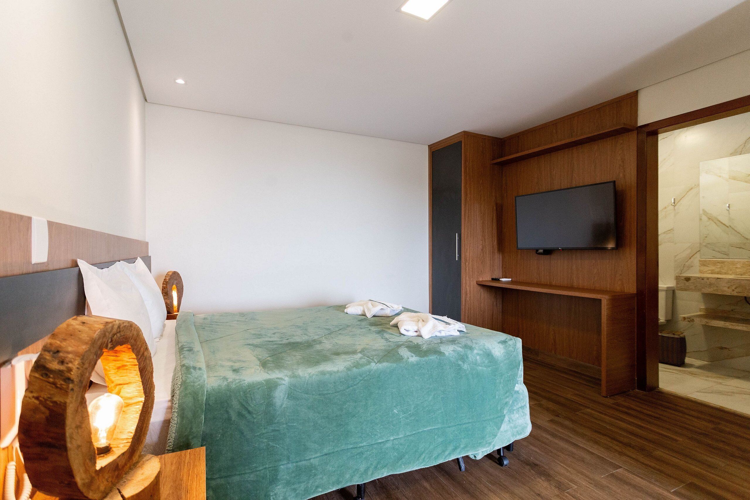 A modern hotel room with a bed, a wooden headboard, a flat-screen TV mounted on the wall, and a bathroom visible through an open door.