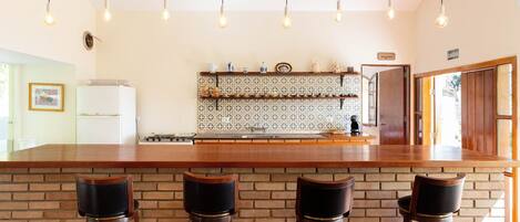 A kitchen with a wooden bar counter, three stools, hanging light bulbs, and a tiled backsplash.
