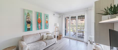 Uma sala de estar moderna com um sofá bege, uma TV de tela plana e uma janela grande com vista para o exterior.