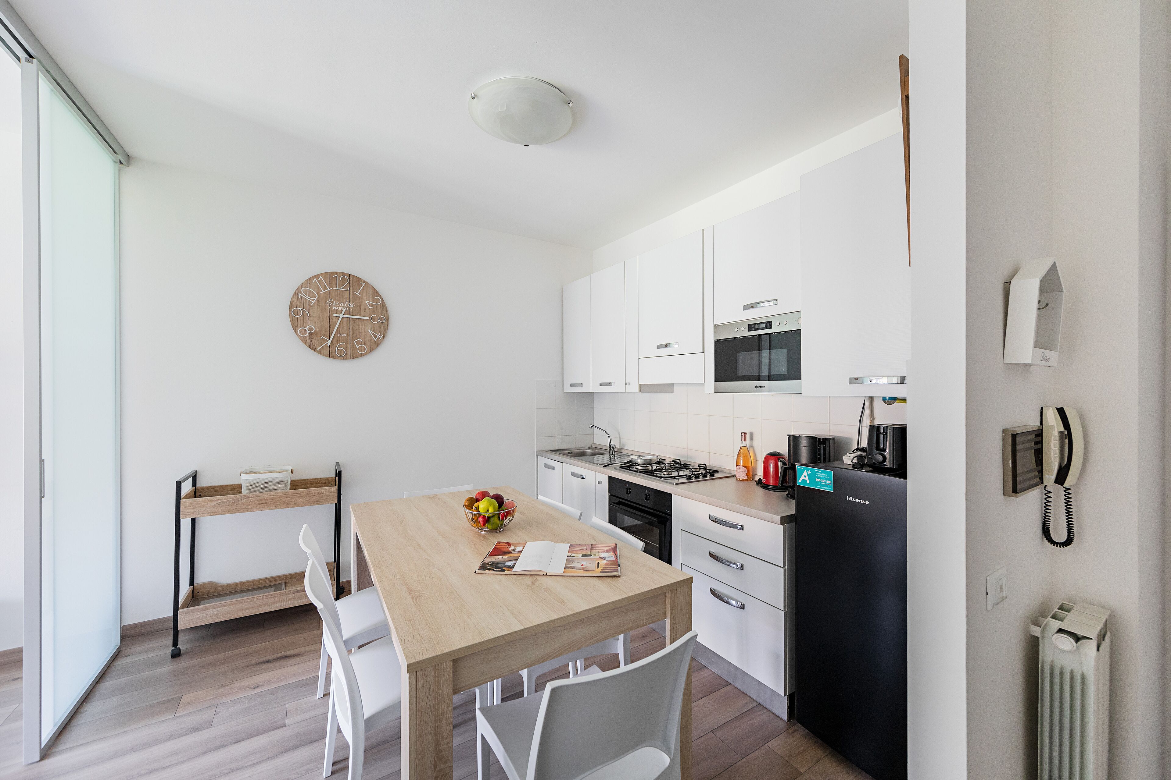 A modern kitchen with white cabinets, a wooden dining table, and a clock on the wall.