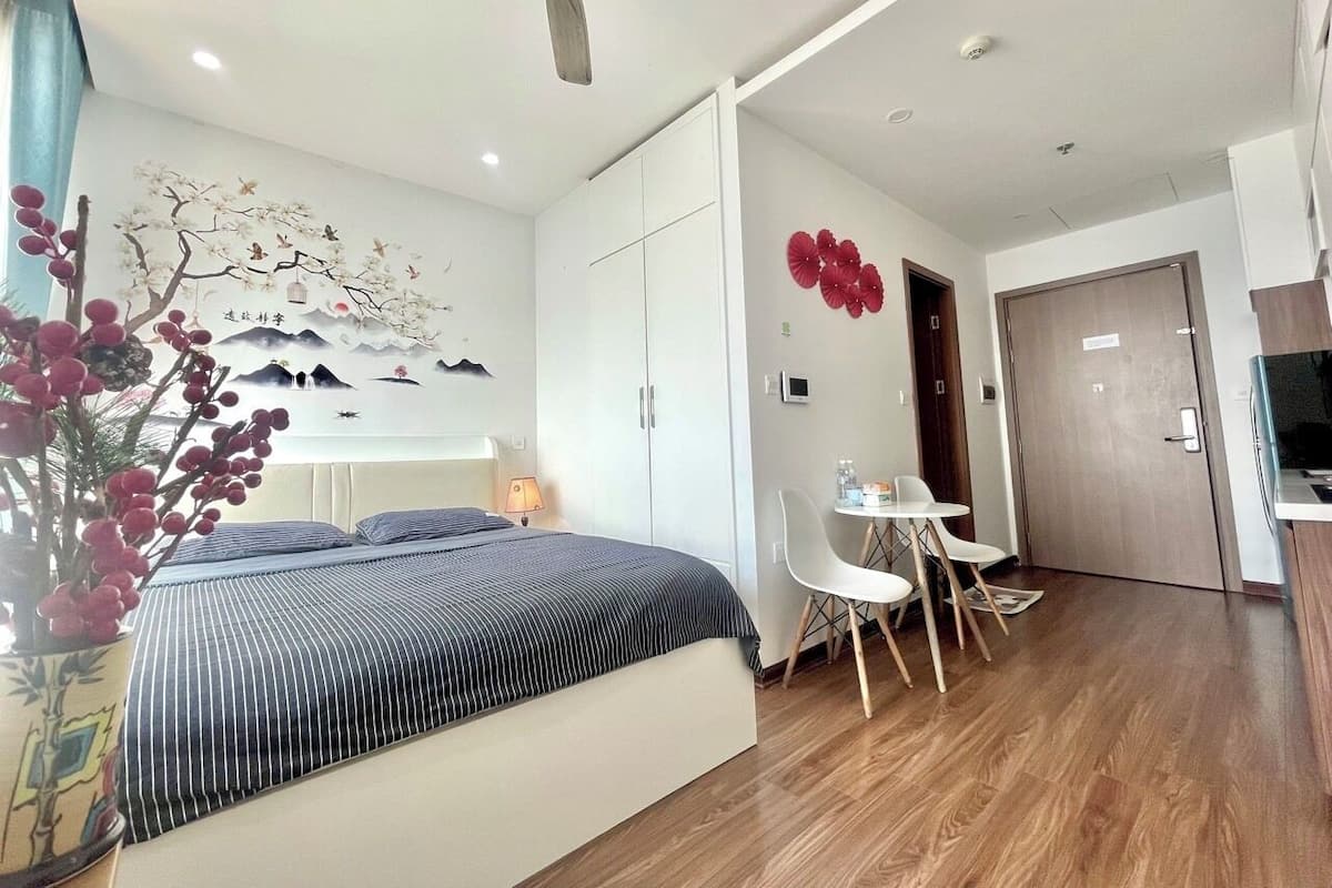 A modern hotel room with a bed, a desk, and a chair. There is a ceiling fan, a wall-mounted TV, and a door leading to another room.