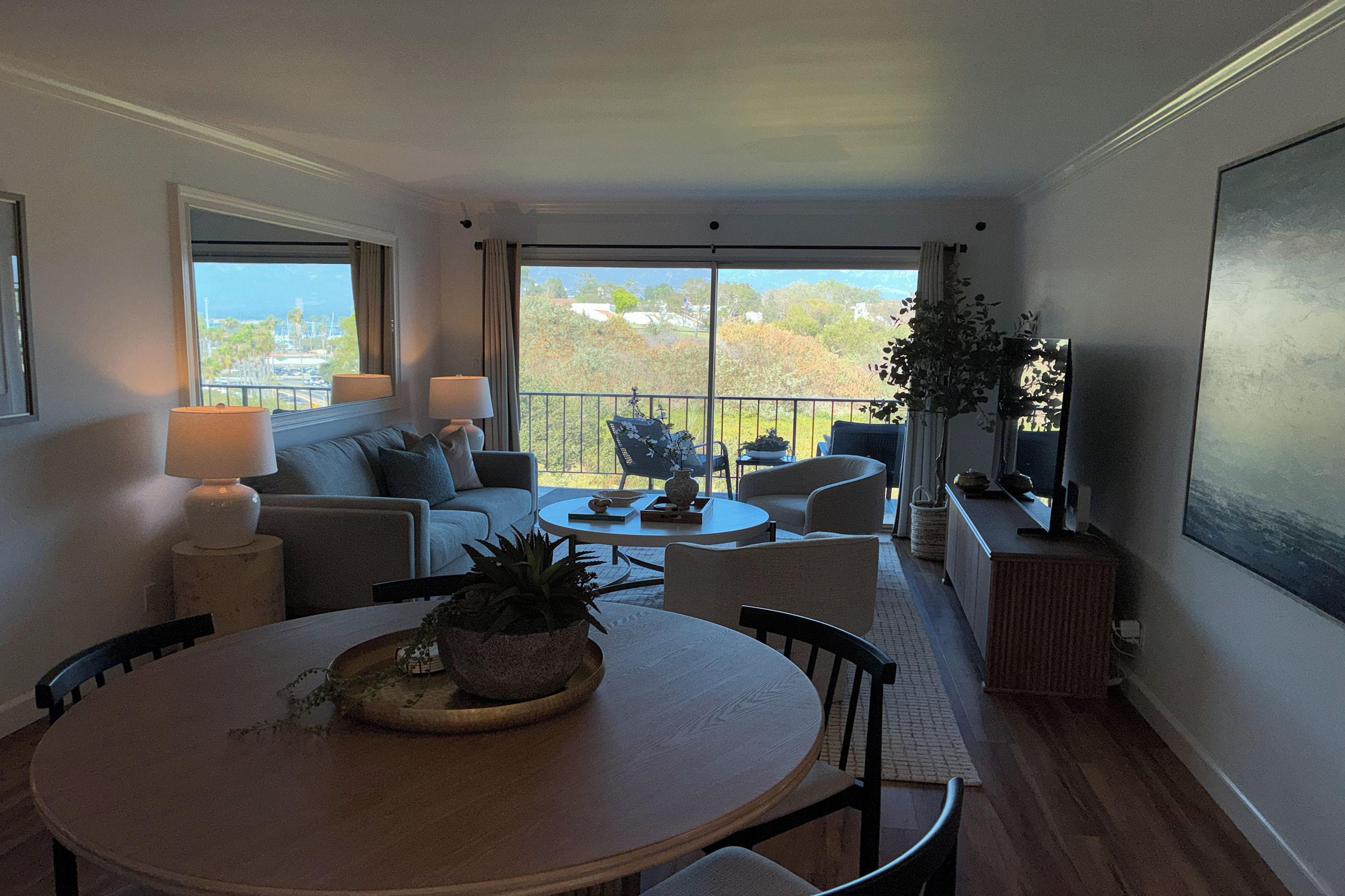 A modern living room with a sofa, two armchairs, a coffee table, and a dining table with chairs. There is a TV on a stand and a large window with a view of trees and buildings.