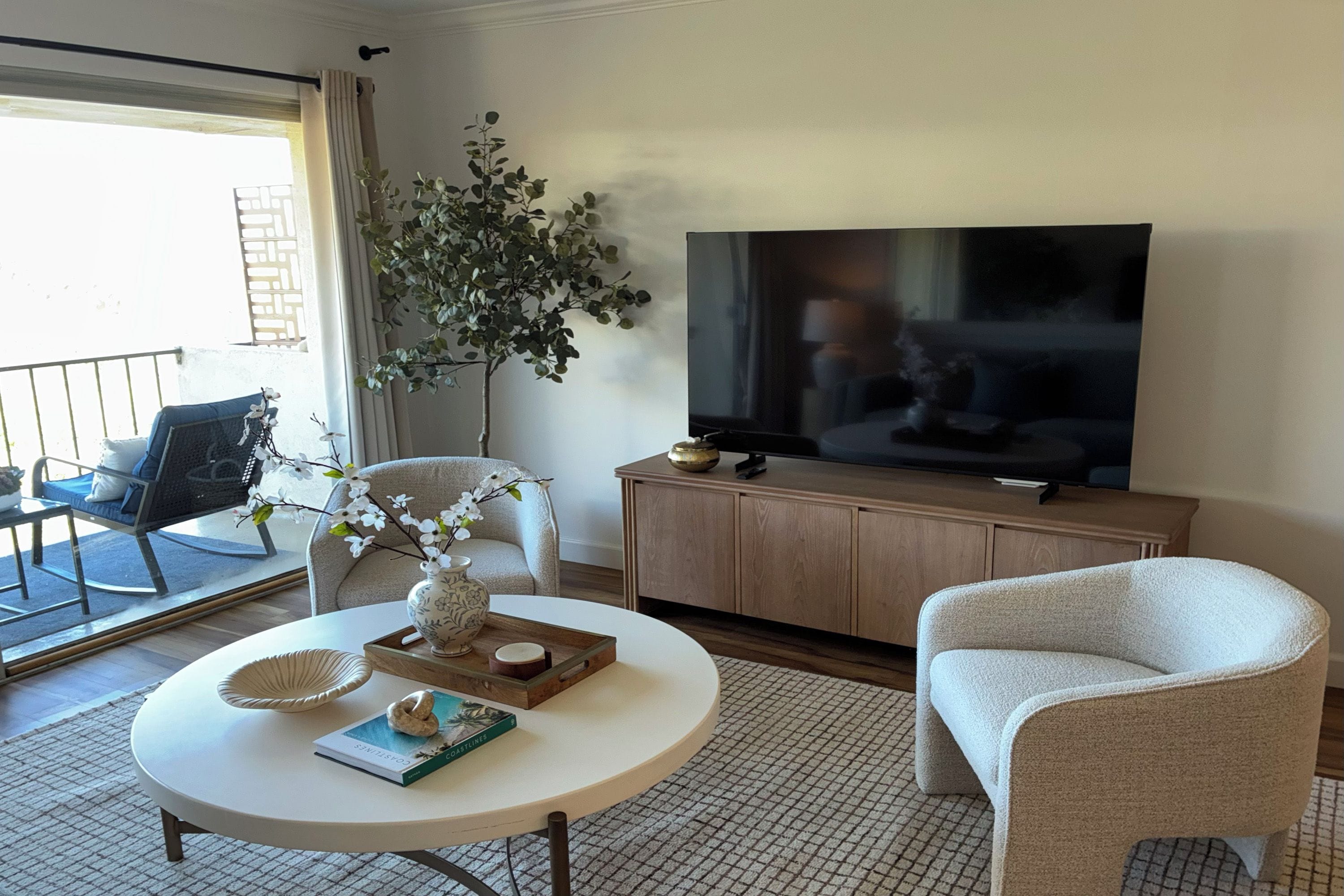 A living room with a round coffee table, a white armchair, a sofa, a television, and a large plant.