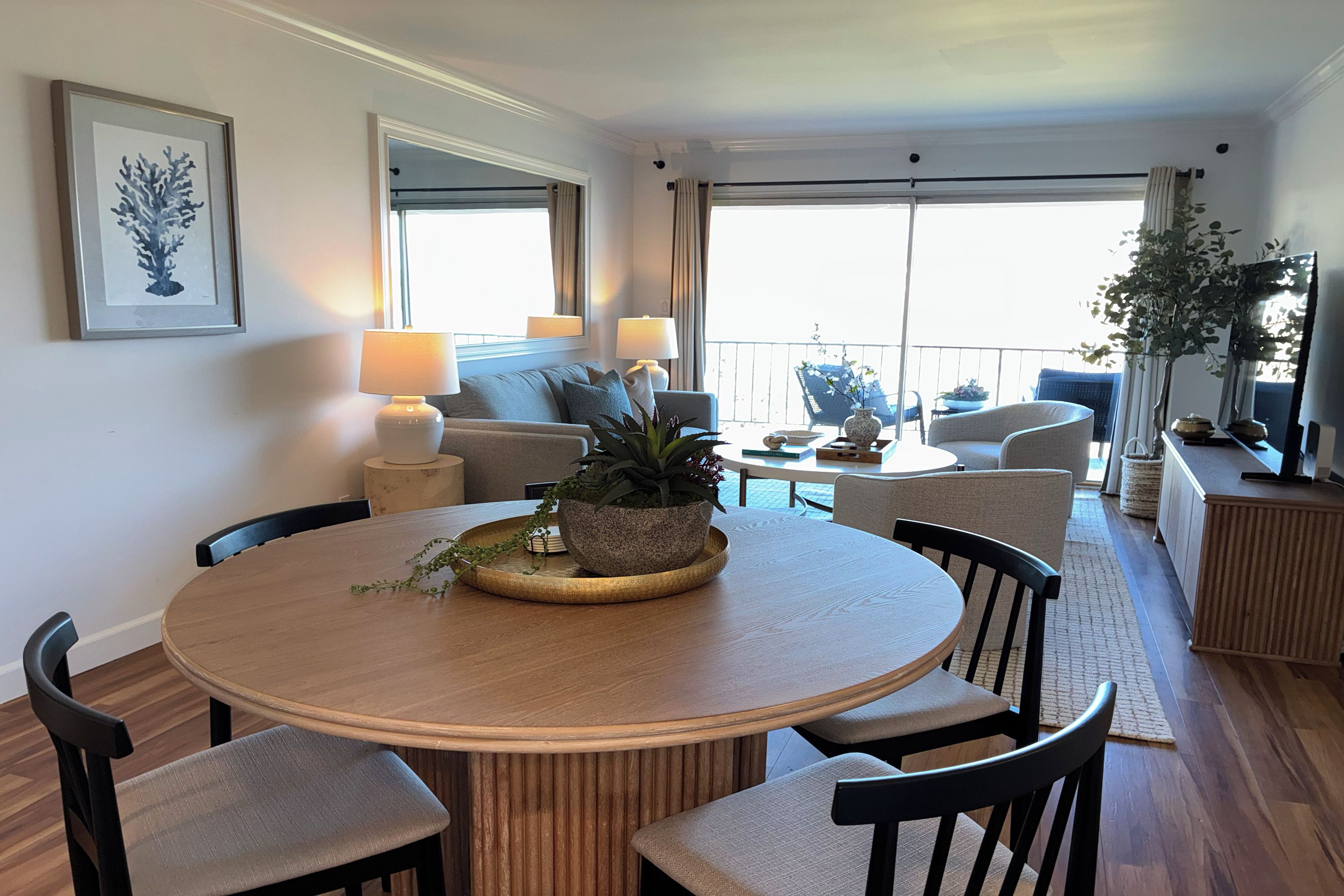 A modern living room with a round dining table, comfortable seating, a large window with curtains, and a balcony with outdoor furniture.