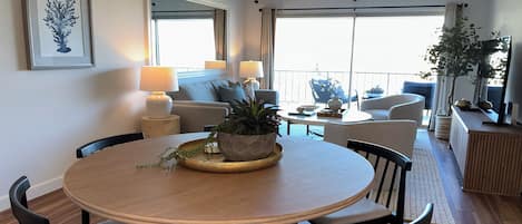 A modern living room with a round dining table, comfortable seating, a large window with curtains, and a balcony with outdoor furniture.