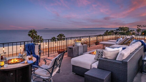 Une terrasse avec des sièges extérieurs, un foyer et une vue sur la plage et l’océan au crépuscule.