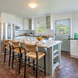 Una cocina con armarios blancos, una isla central con taburetes y vistas a través de una ventana, además de una puerta con vistas al exterior.