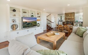 Una sala de estar moderna con un televisor grande, muebles blancos, una chimenea de piedra y un comedor con sillas de mimbre.