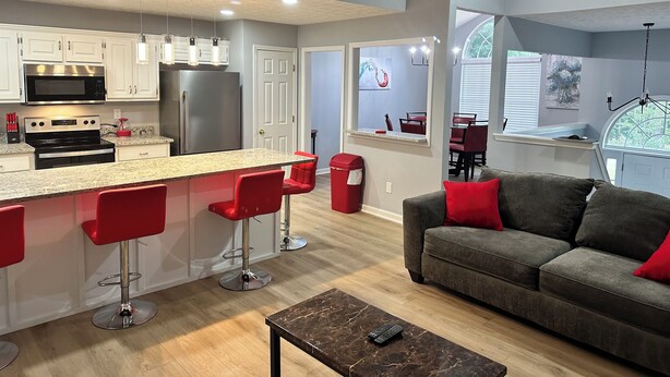 A modern kitchen with a central island, red bar stools, a grey sofa with red cushions, and a dining area with a view through a window. - Charming Locust Grove GetAwayHome 4bd/3bath (Locust Grove)