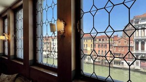Ein Zimmer mit großen Fenstern, die geometrische Metallgitter aufweisen, einem Holzboden und Blick auf historische Gebäude an einem Kanal.