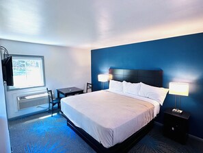A hotel room with a bed, bedside lamps, a desk, and a chair. - Budget Host Inn Manistique (Manistique)