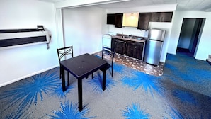 Una cocina compacta con estufa, pileta y refrigerador, una mesa de comedor con sillas y una alfombra estampada.