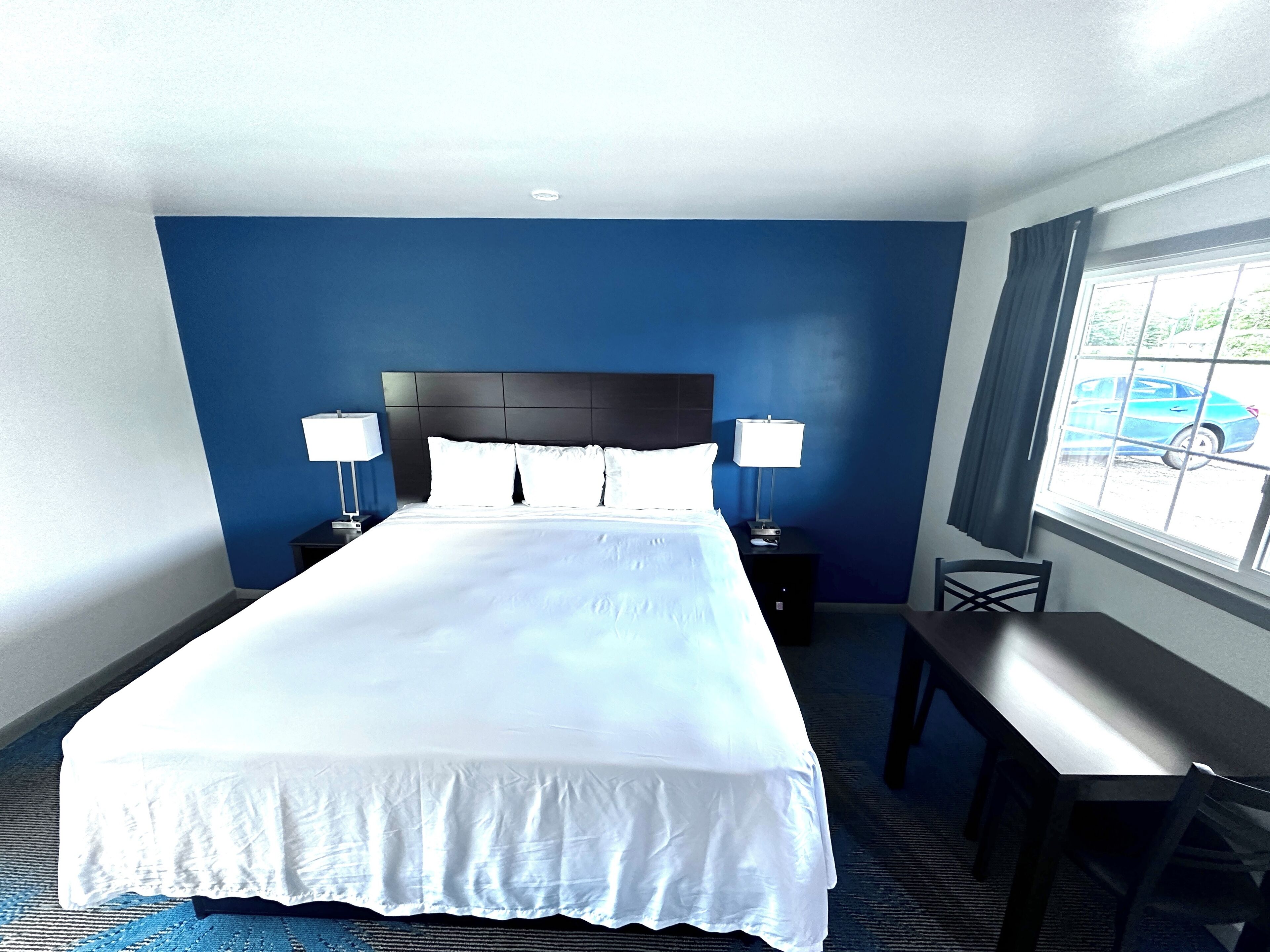 A hotel room with a large bed, two bedside tables with lamps, a desk, and a window with a view of a car.