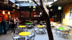 An indoor dining area with multiple tables and chairs, a central tree, and a bar area in the background. - GoodMo Budapest Smart Rooms (Budapest)