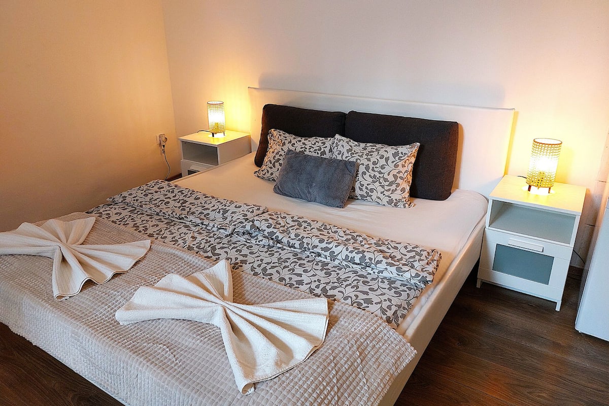 A neatly made bed with a patterned bedspread and two decorative towels, two bedside lamps, and a nightstand with a lamp.