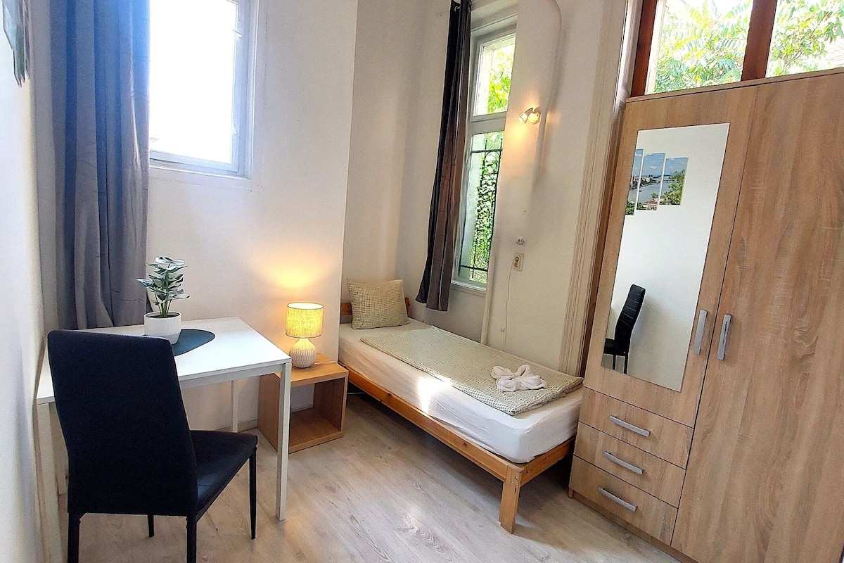 A small, well-lit room with a single bed, a desk with a chair, a lamp, and a wardrobe.